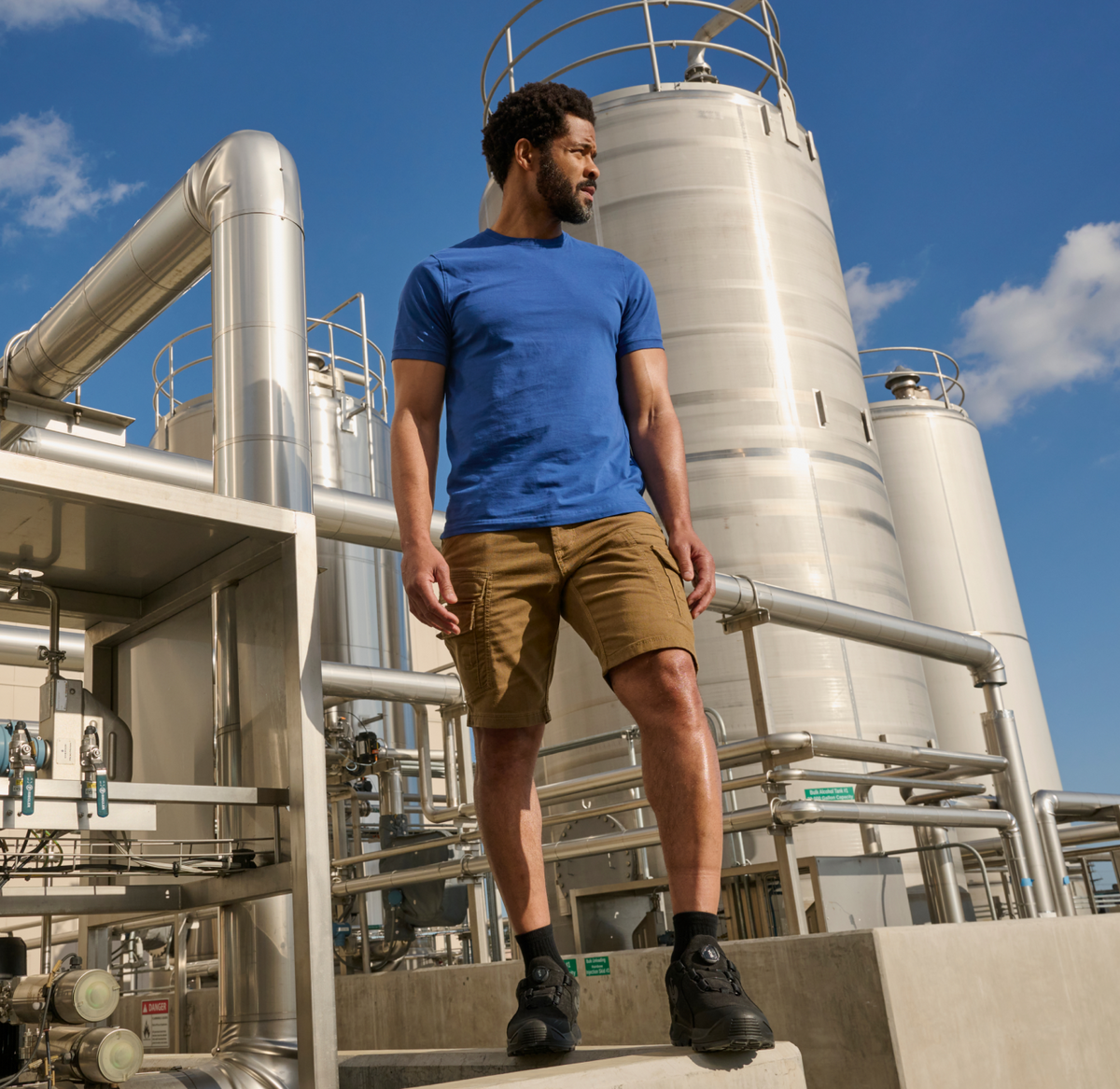 A man in a blue shirt and brown shorts stands confidently among large industrial tanks and pipes, looking to the side under a clear blue sky.