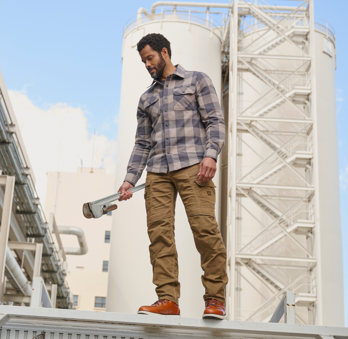A man in work clothes holds a large pipe wrench while standing outdoors near industrial storage tanks and pipes, looking down.