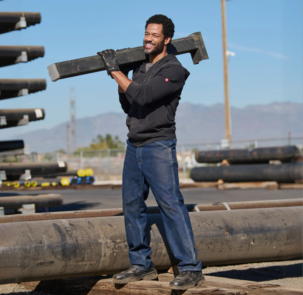 A man in jeans and a black hoodie smiles while carrying a large metal beam on his shoulder at an industrial outdoor worksite with pipes and mountains in the background.