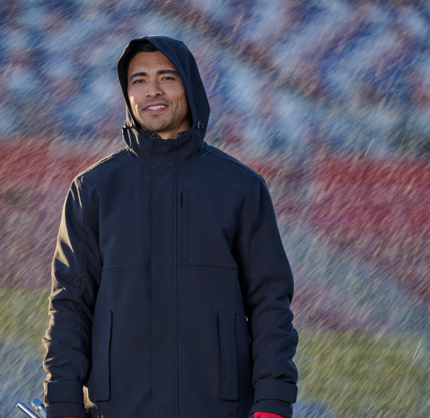 A man in a hooded black jacket stands outdoors in the rain, smiling slightly. The background is blurred with red and blue colors. He holds something silver in his left hand.