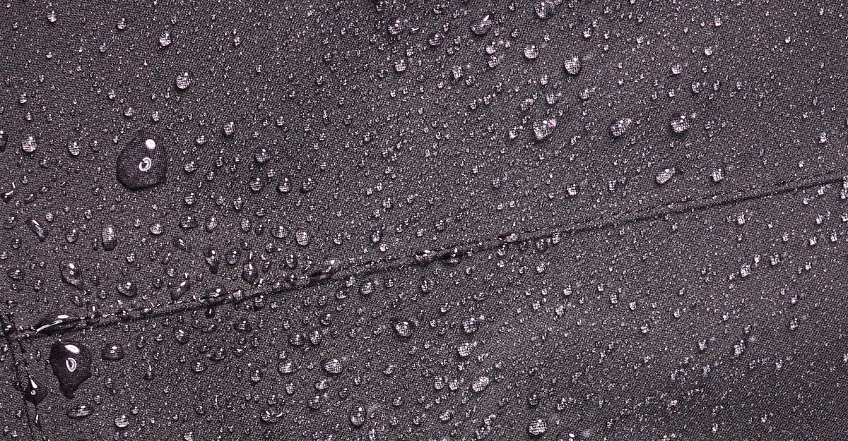 Close-up of water droplets on a dark, textured fabric surface, showing the materials water-resistant or waterproof properties.