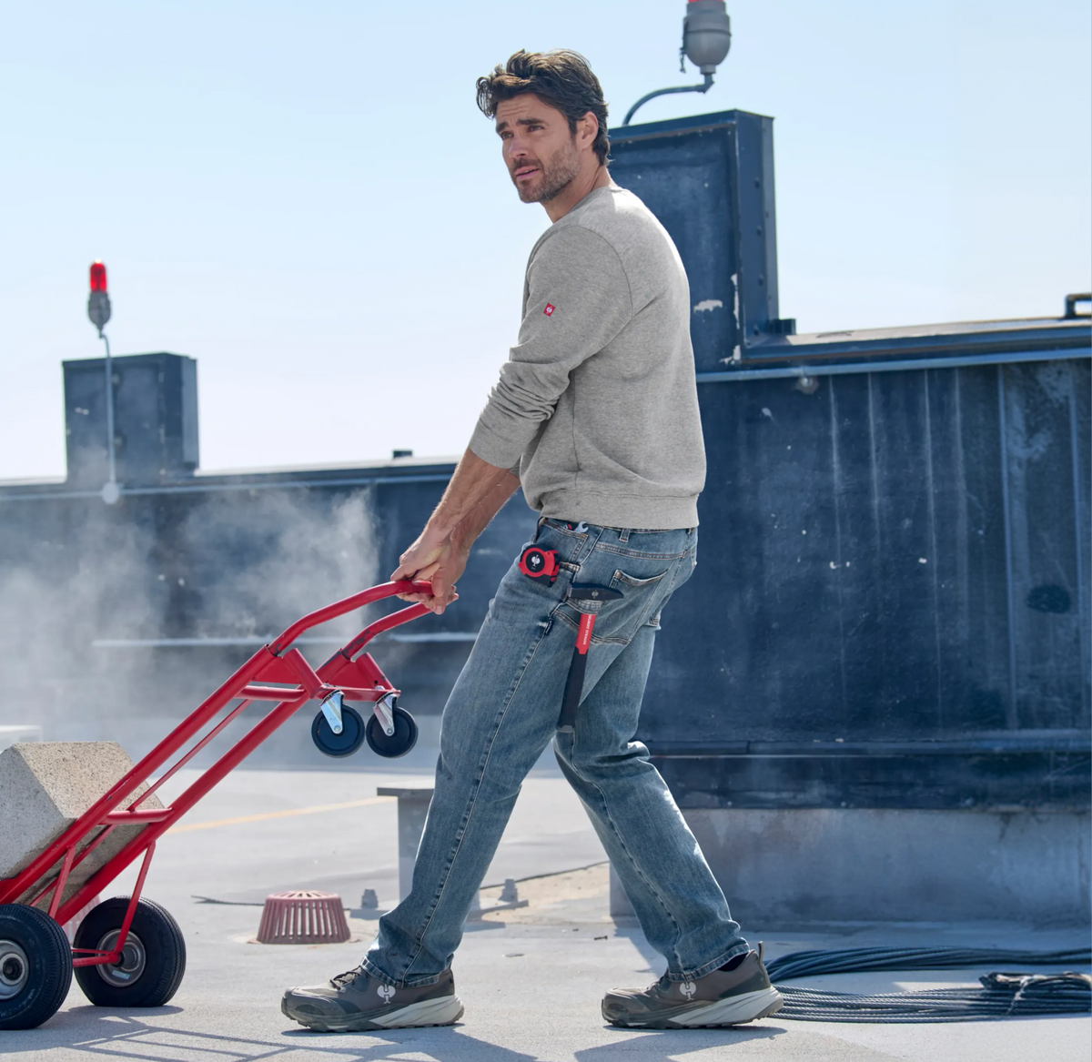 A man in a gray sweatshirt and jeans uses a red hand truck to move a concrete block outdoors. Tools hang from his jeans, and steam or smoke rises in the background. Industrial equipment is visible behind him.