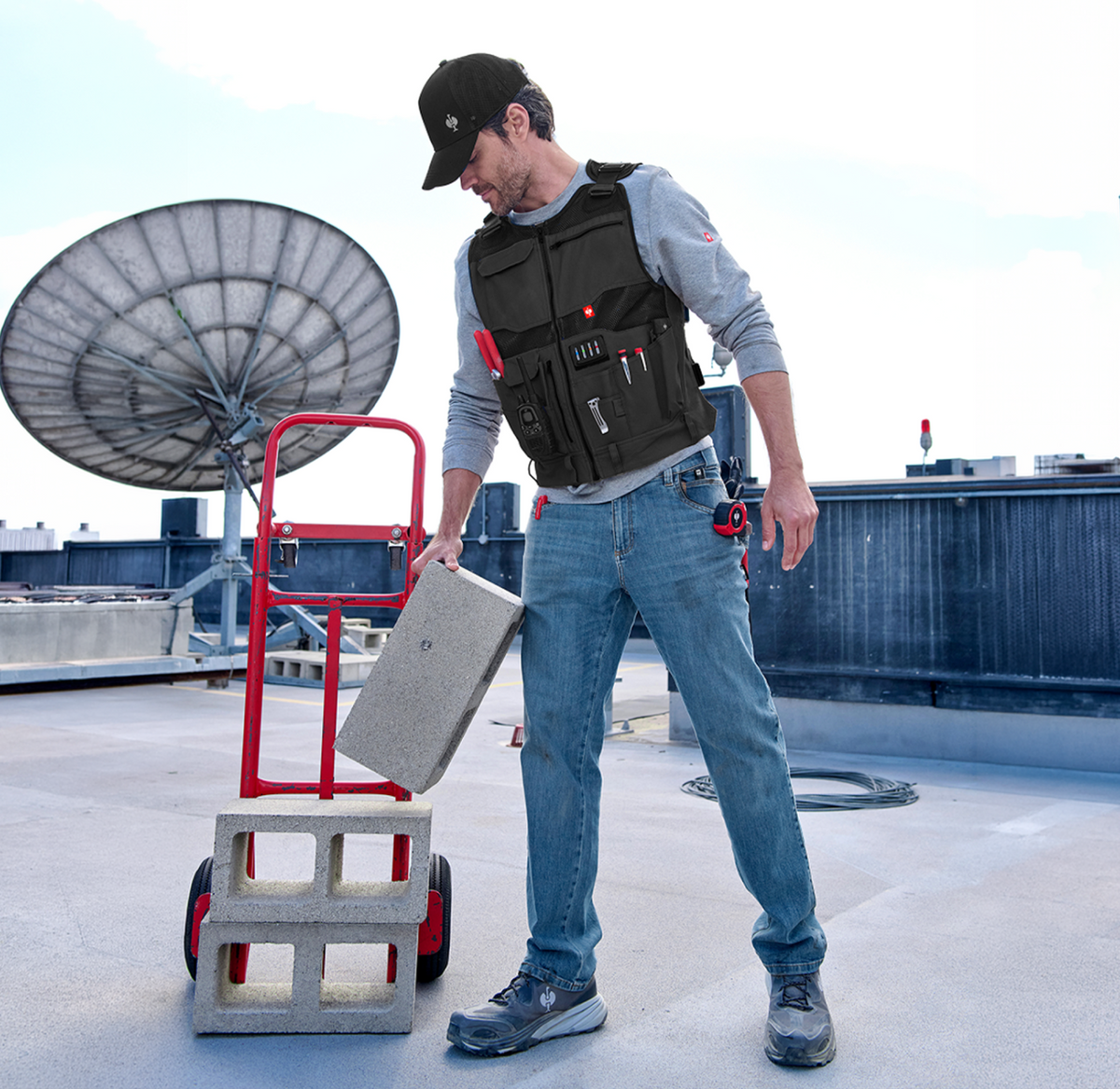 A man in a black vest and cap lifts a concrete block onto a red hand truck on a rooftop, with a large satellite dish and blue industrial structures in the background.