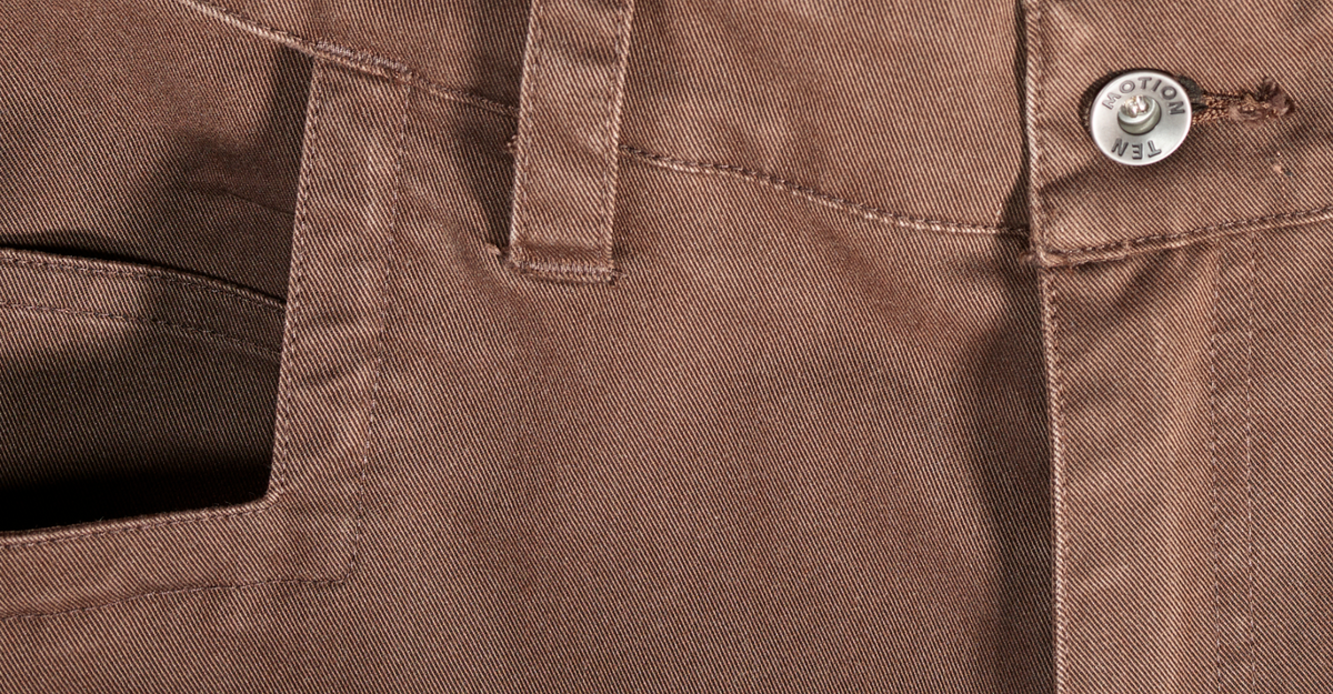 Close-up of brown denim pants showing front pocket, belt loops, and a silver Motion Ten branded button at the waistband.
