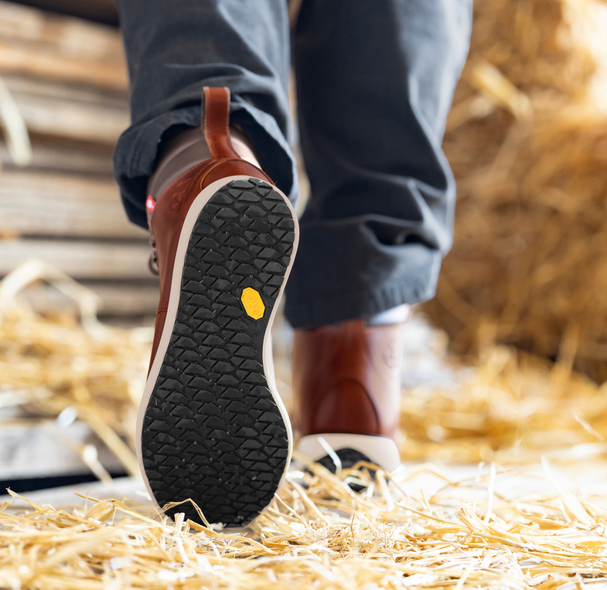 A close-up of a person in brown shoes and dark pants walking on straw, focusing on the textured sole of the shoe with a yellow logo. Wooden planks and hay are in the background.