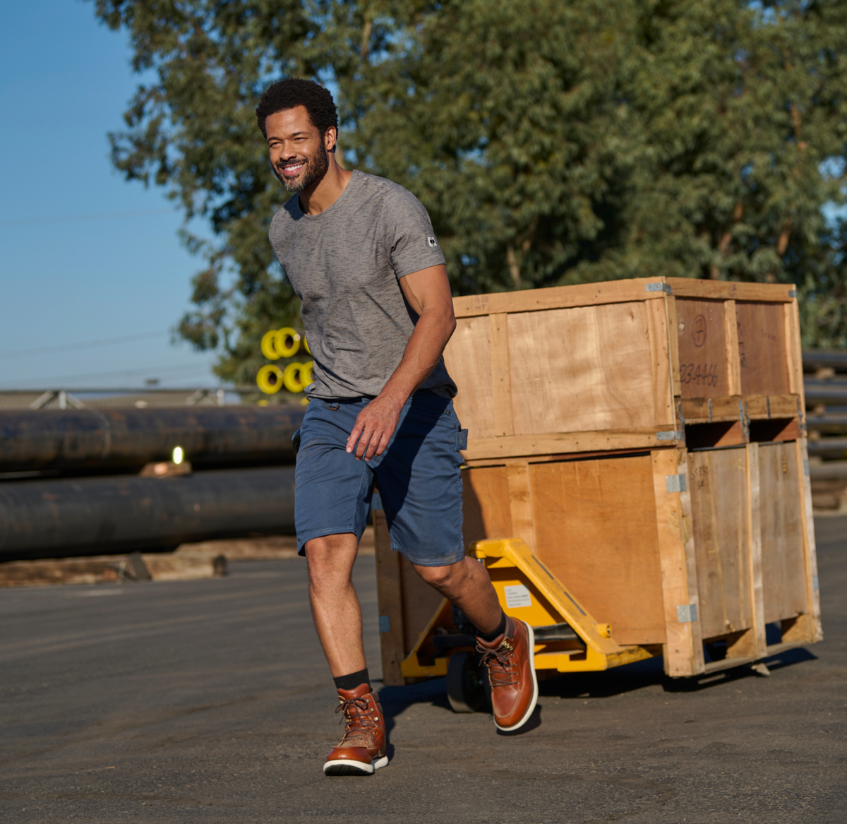 A man in a gray t-shirt and blue shorts smiles while pulling a wooden crate on a yellow hand truck outdoors, with large pipes and trees in the background.