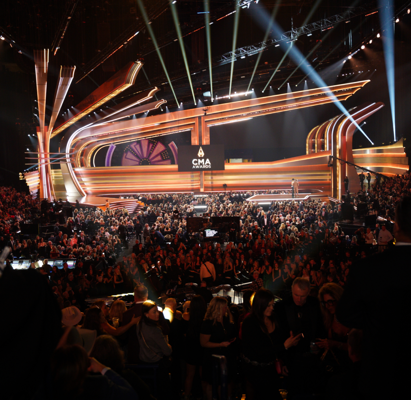 A large crowd fills an arena facing an elaborate, brightly lit stage with CMA Awards displayed on a central screen. The atmosphere is lively, with people seated and awaiting the event.