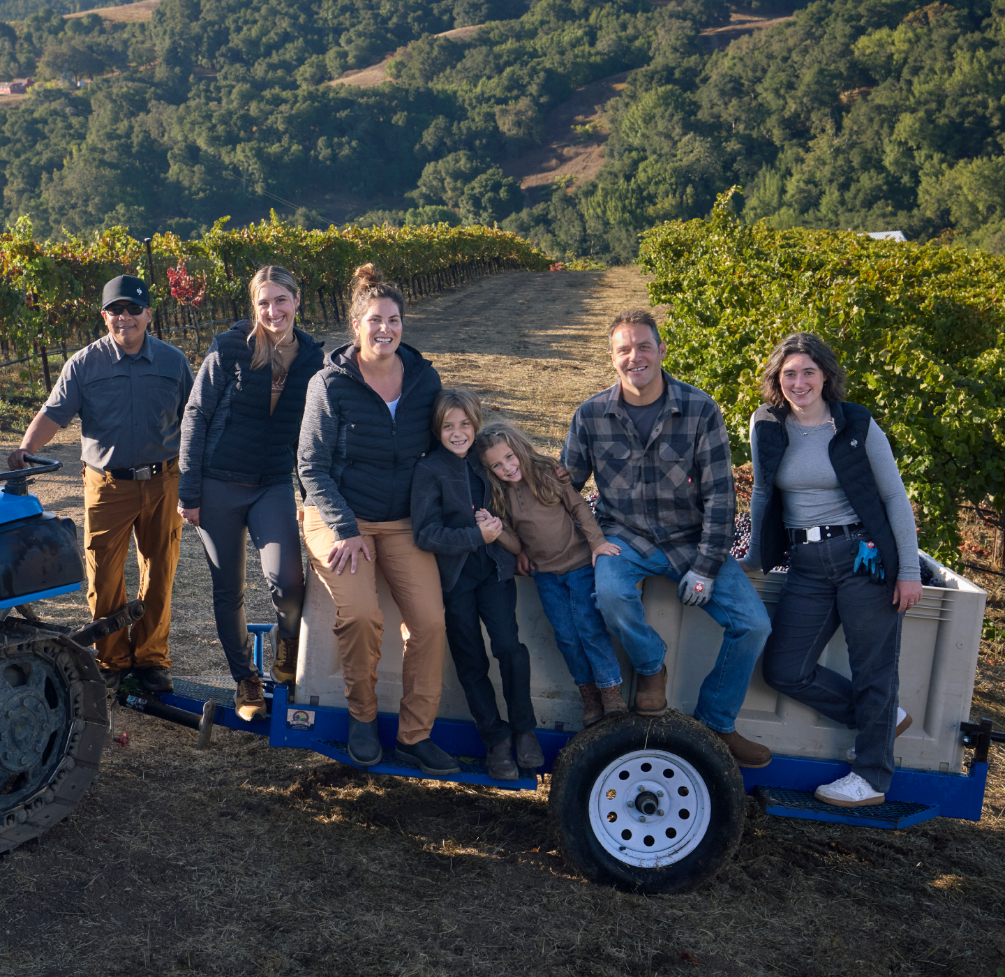 Seven people, including two children, smile and pose on and around a small trailer attached to a tractor in a vineyard, with rows of grapevines and forested hills in the background.