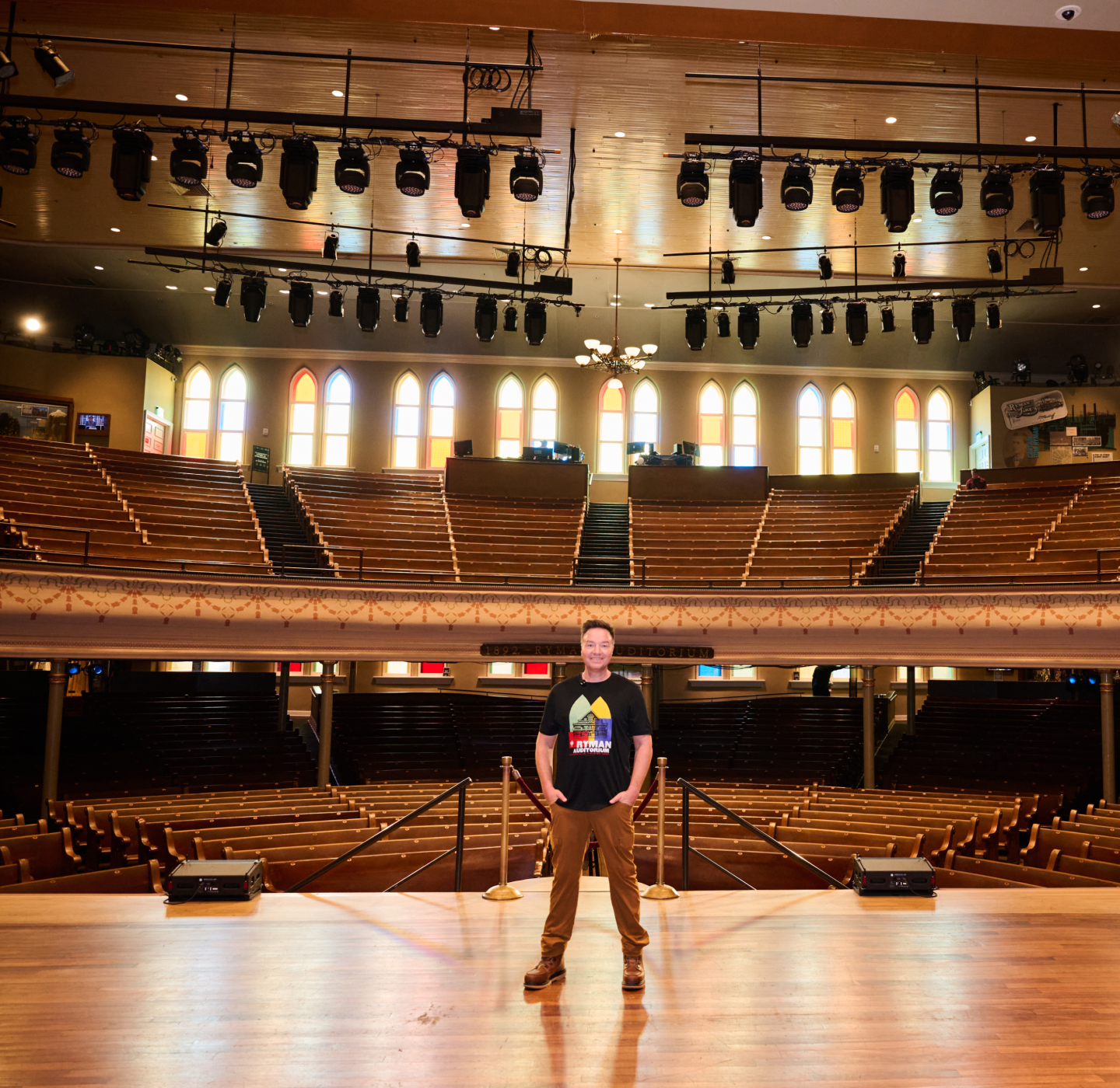A person stands center stage in a large, empty theater with wooden seats and arched stained-glass windows. Stage lights hang above and rows of balcony seating are visible in the background.