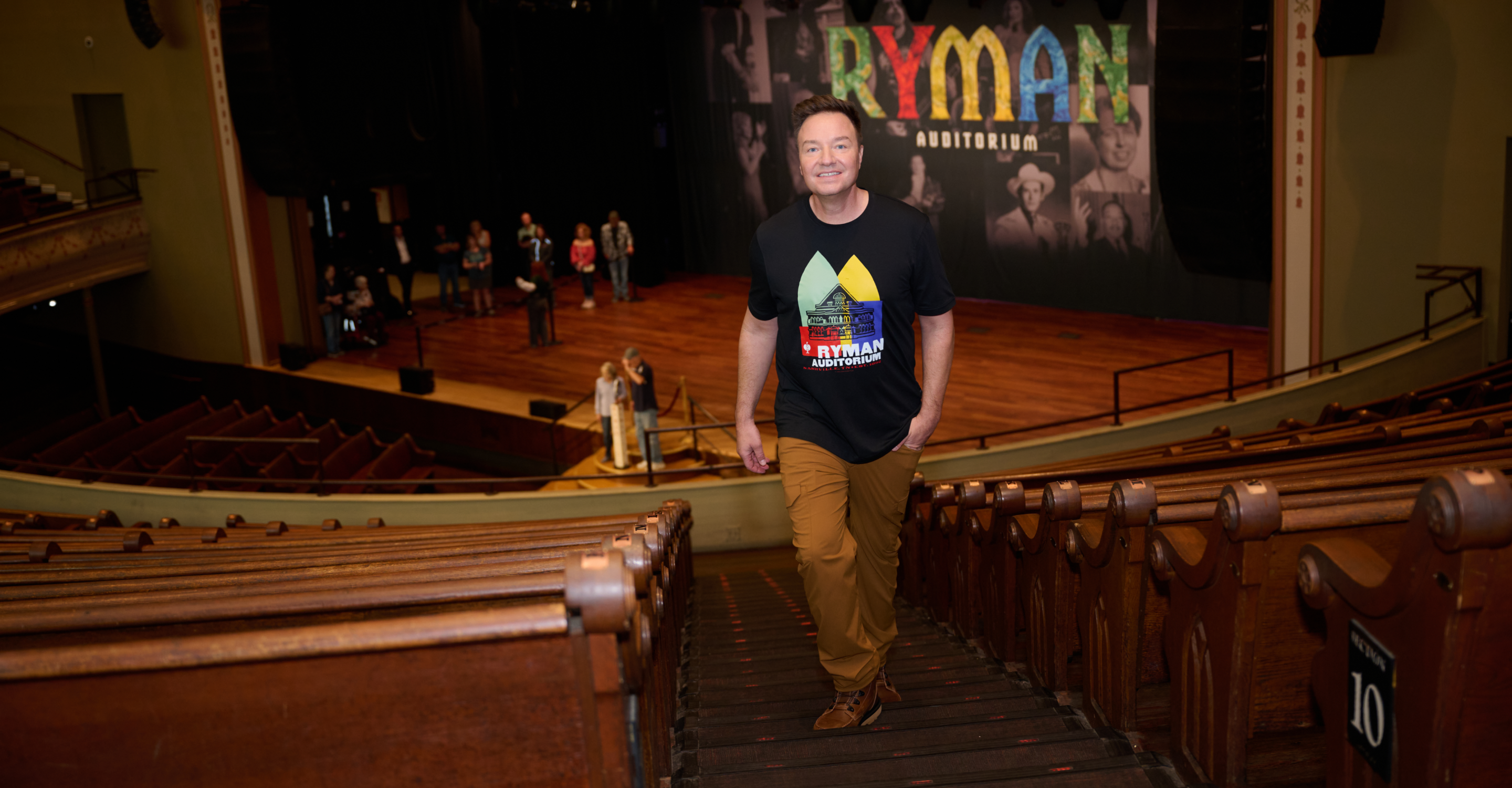 A man in a Ryman Auditorium T-shirt smiles while walking up auditorium stairs, with a stage and several people visible in the background.