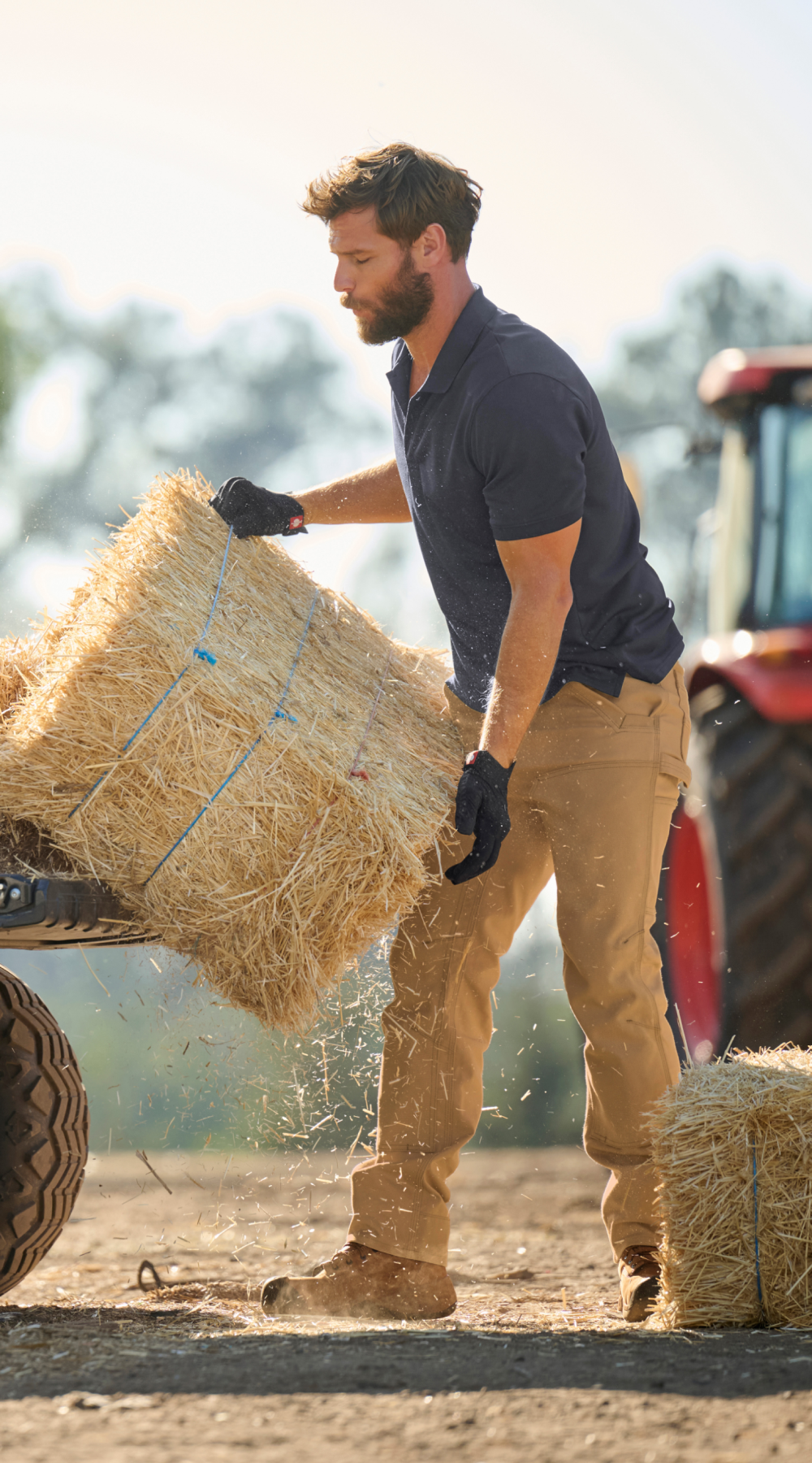 A man wearing gloves and work clothes lifts a bale of hay onto a trailer in a rural outdoor setting, with a red tractor visible in the background.