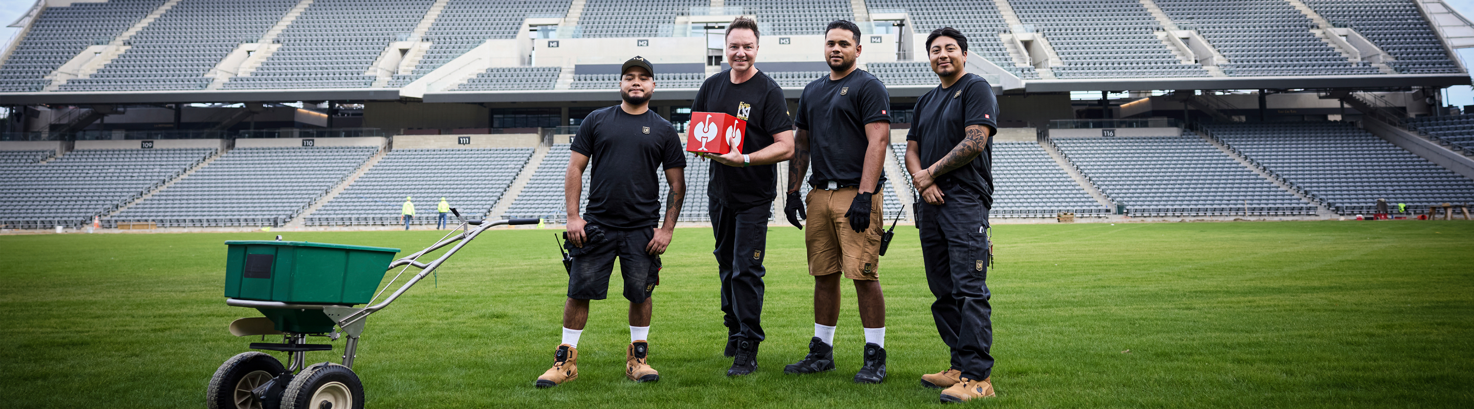 Four men in work uniforms stand on a stadium field, smiling at the camera. One holds a red first aid kit. A green utility cart is nearby. The stadium seats are empty in the background.
