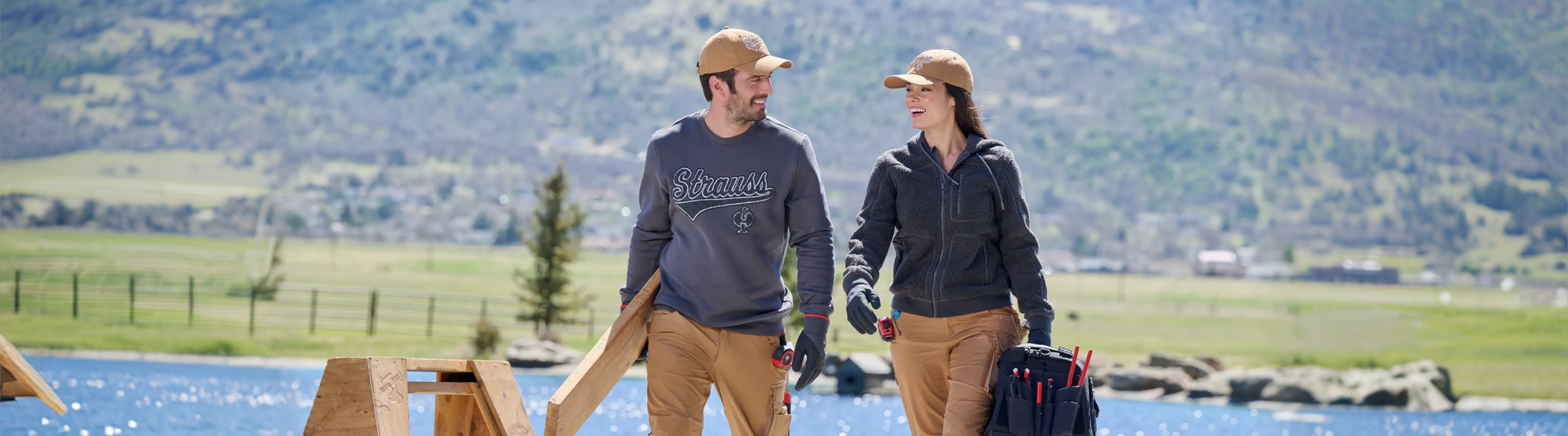 Two people in work clothes and hats smile at each other while carrying tools outdoors near a lake, with mountains and green fields in the background.