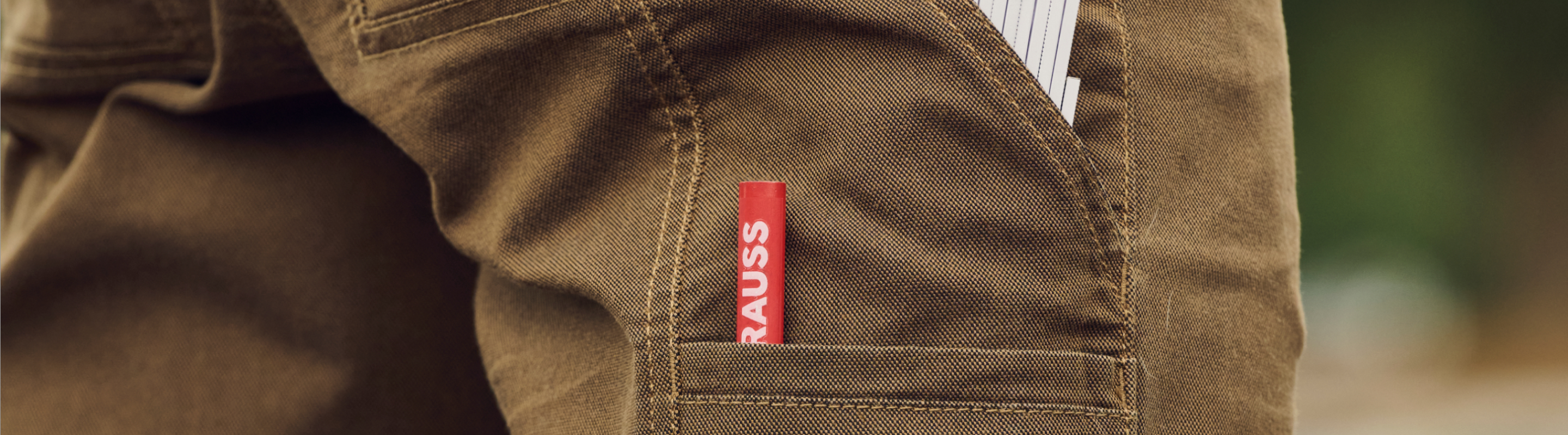 Close-up of a brown pants pocket holding a red pencil labeled RAUSS and a folded white paper peeking out from the top of the pocket.