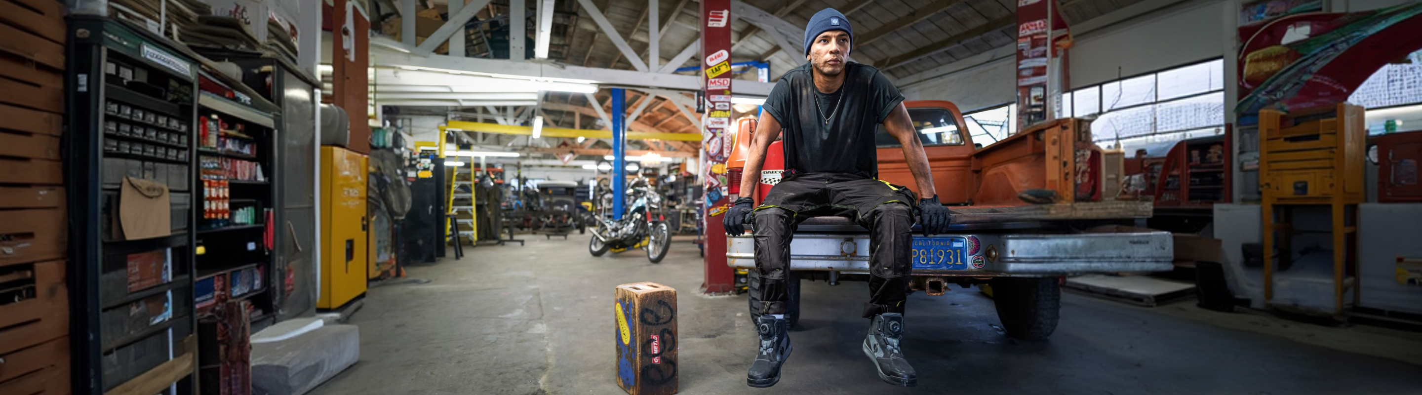 A person in work boots and gloves sits on the tailgate of a pickup truck inside a busy auto repair shop, surrounded by tools, equipment, and vehicles.