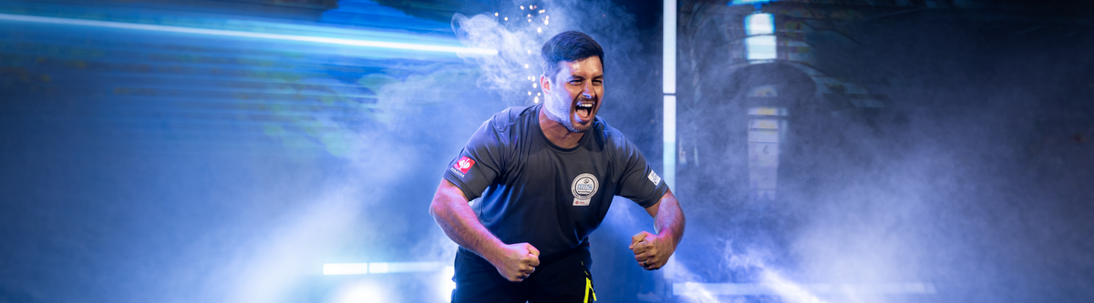 A man in a gray shirt with an HVAC logo is shouting energetically with clenched fists. Bright lights and smoke create a dramatic background.
