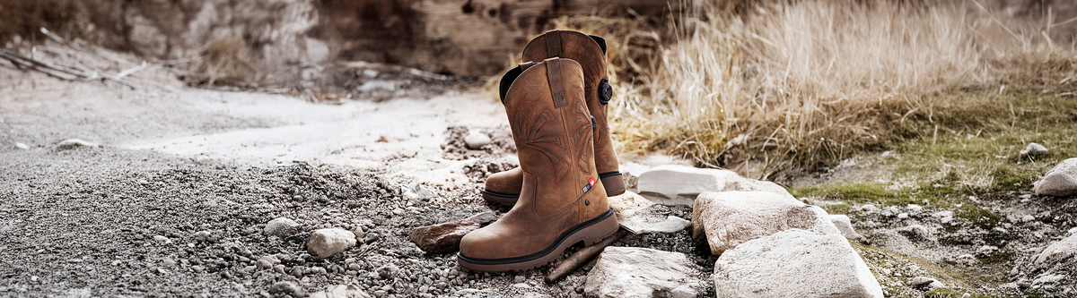 A pair of brown leather work boots stands on rocky ground outdoors, with dry grass and stones in the background.