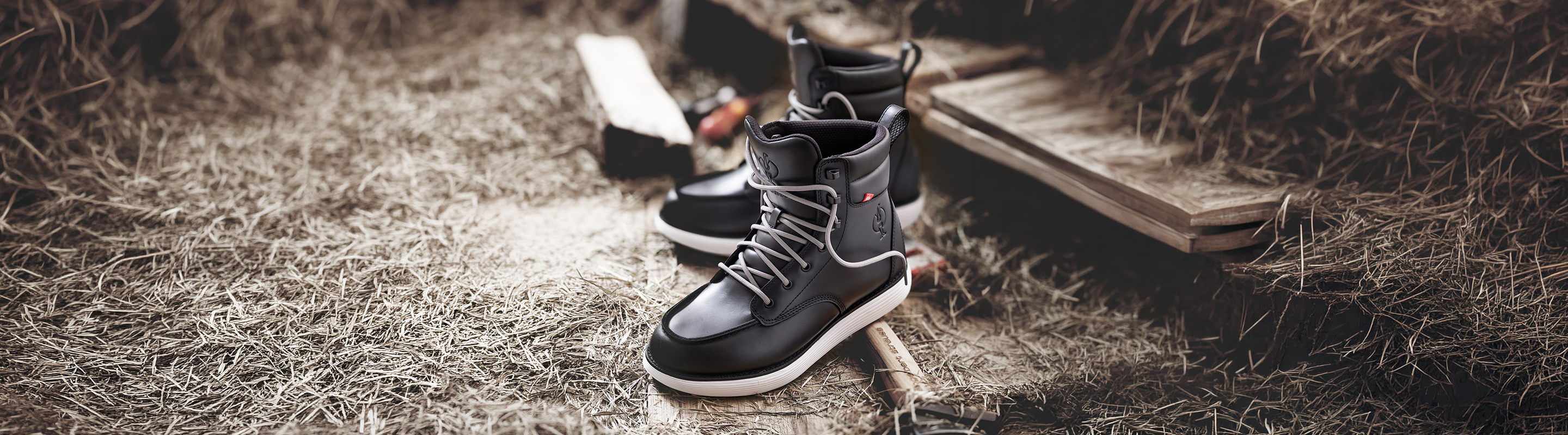 A pair of black high-top sneakers with white soles is placed on dry grass next to a wooden step and a garden tool, suggesting an outdoor rustic setting.