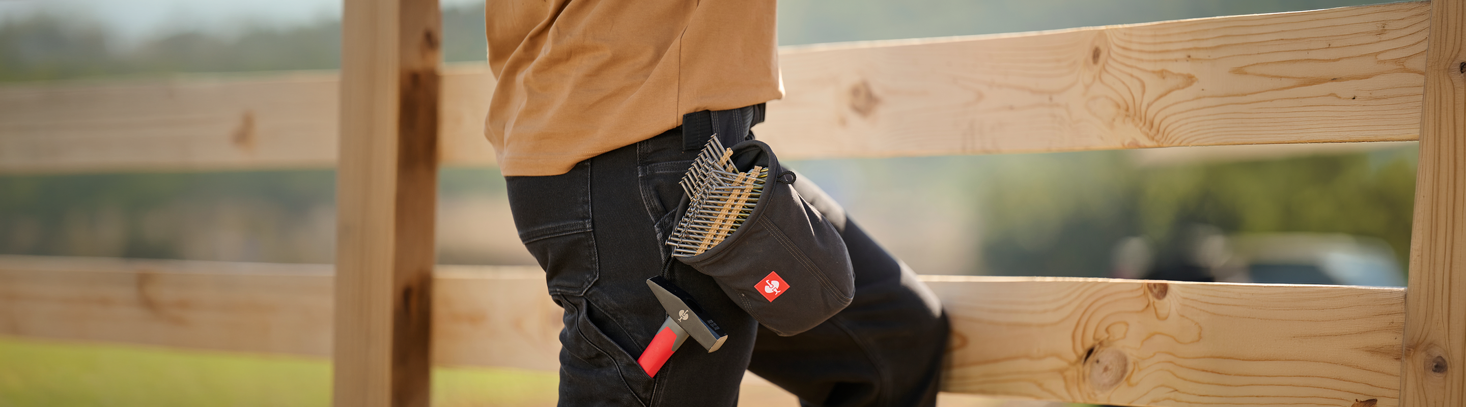 A person in dark jeans and a tan shirt stands by a wooden fence, holding a tool belt filled with nails and a hammer. The background is blurred greenery.