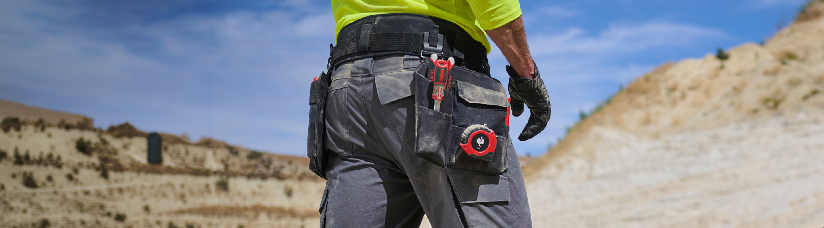 A person wearing a utility belt with tools stands outdoors in a rocky, desert-like area, facing away from the camera. The persons bright shirt and gloves suggest construction or surveying work.