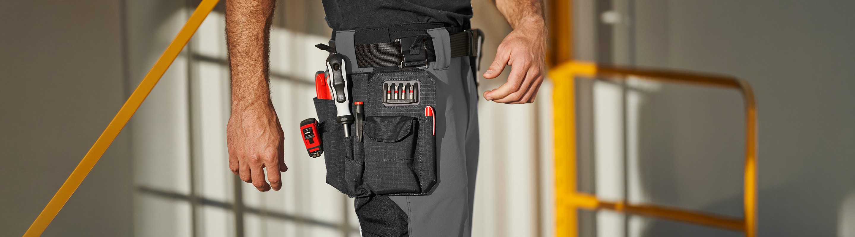 A person wearing gray work pants with a tool belt holding a tape measure, screwdriver, pliers, pen, and a set of bits. The background shows blurred metal railings and walls.