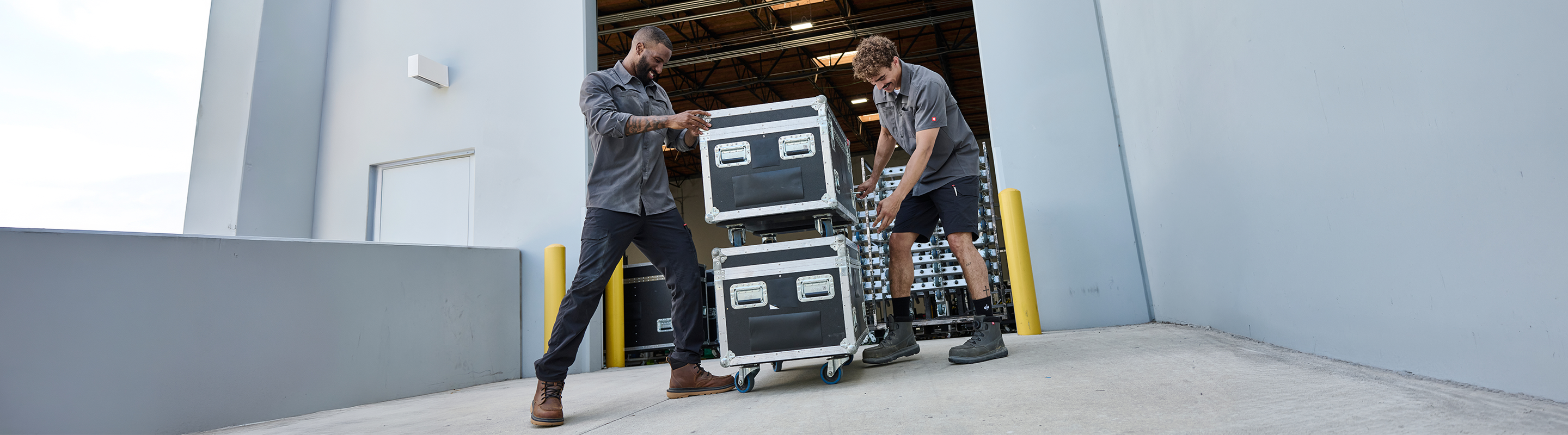 Two people wearing uniforms move large black equipment cases on a dolly down a ramp outside a building, next to an open loading dock door.
