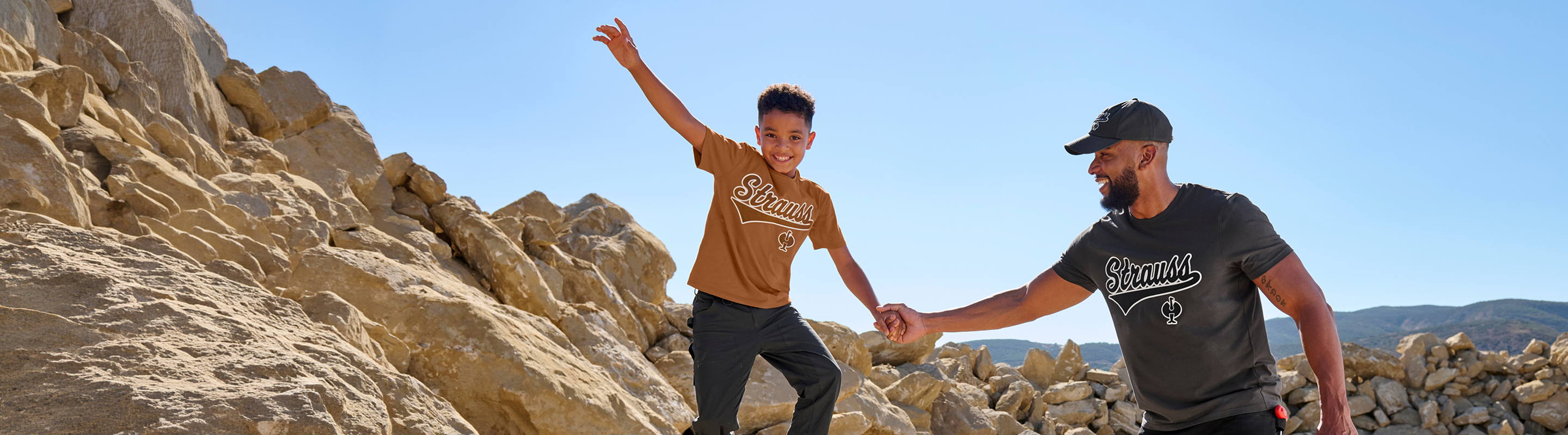 A smiling boy in a brown Skimm shirt climbs rocks, holding hands with a man in a black Skimm shirt. Theyre outdoors with blue sky and rocky hills in the background.