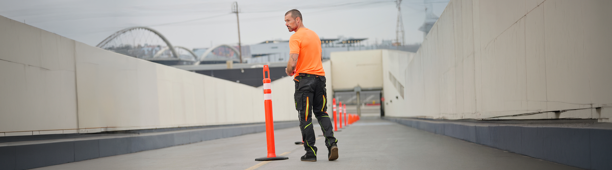 A construction worker in an orange shirt and black pants adjusts an orange traffic cone on an empty roadway, with bridges and buildings visible in the background.