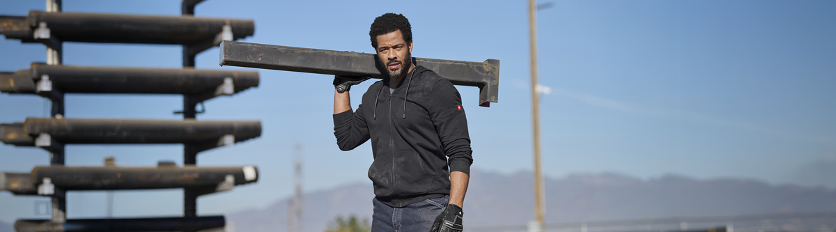 A man in a black hoodie carries a large metal beam on his shoulder at a construction site, with stacks of pipes and mountains in the background under a clear blue sky.