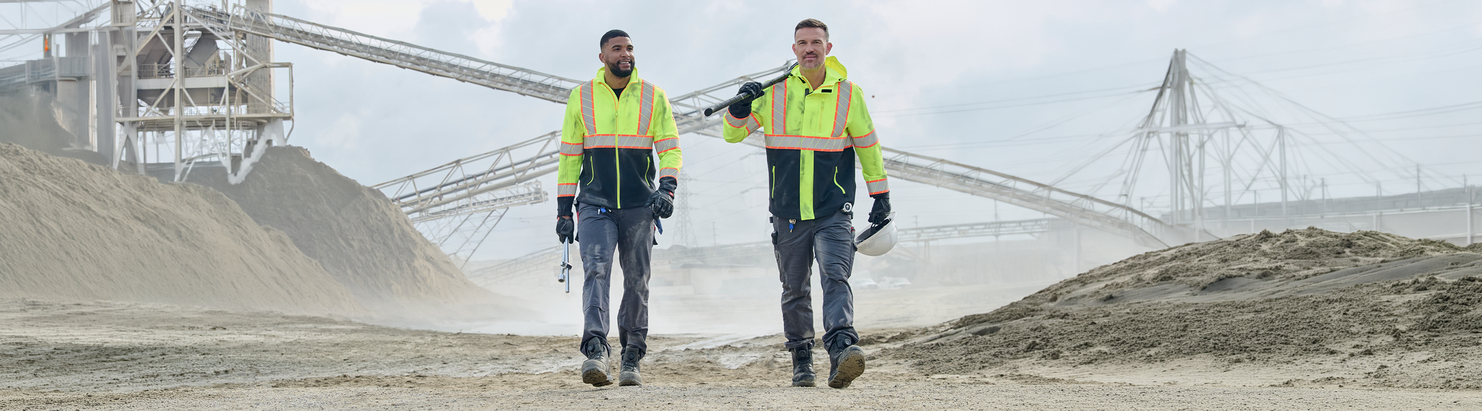 Two workers in high-visibility neon yellow safety vests and dark work clothes walking toward the camera in an industrial quarry or mining site with conveyor belts and piles of material in the background.