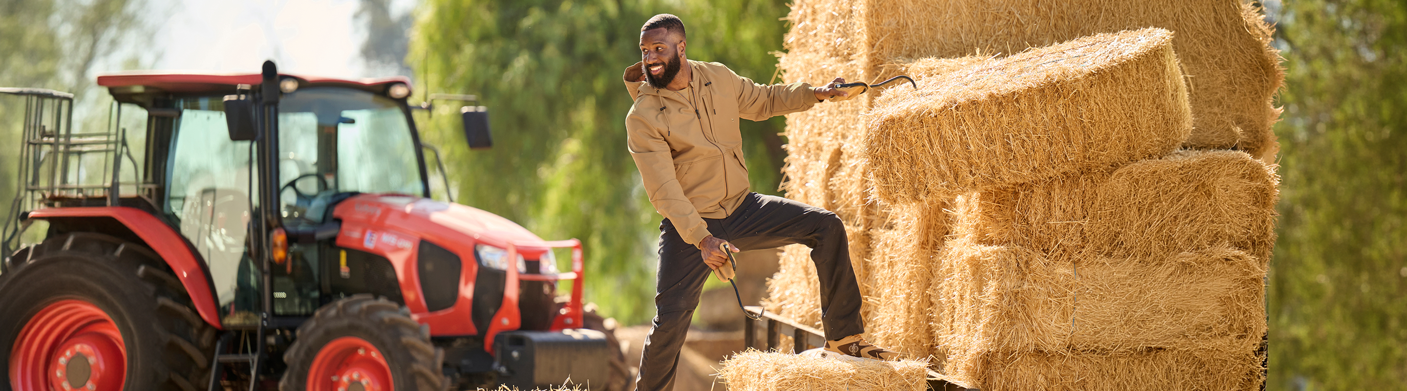 A man in a tan work jacket and dark pants stands on stacked hay bales next to a red tractor, holding a tool in an outdoor farm setting with trees in the background.