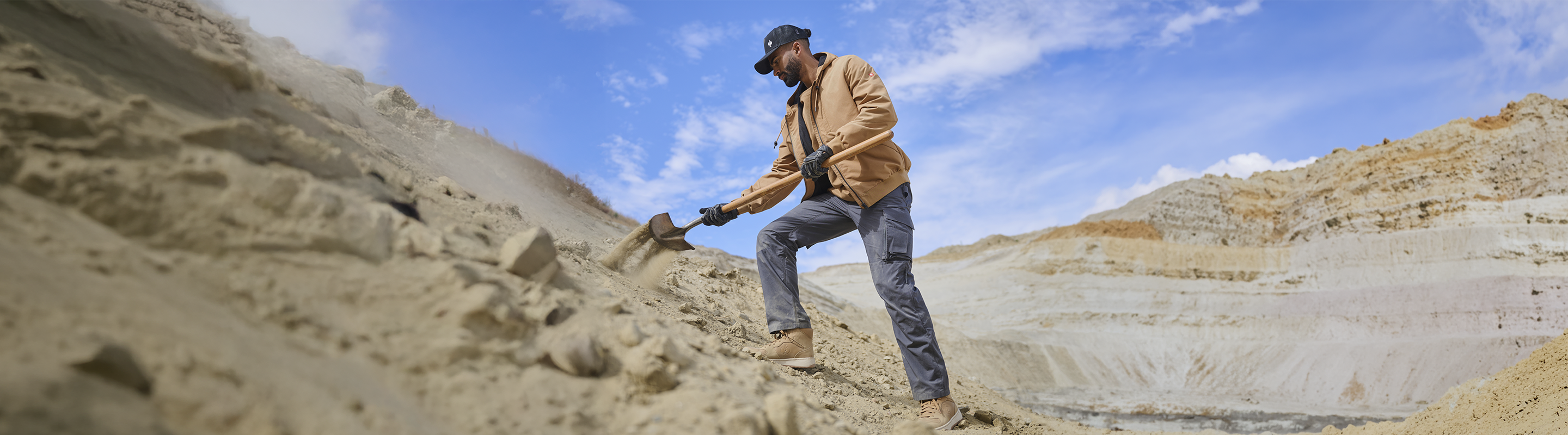 A man wearing a tan jacket, blue jeans, and boots stands in a large open-pit mine, holding a shovel on a rocky slope under a blue sky.