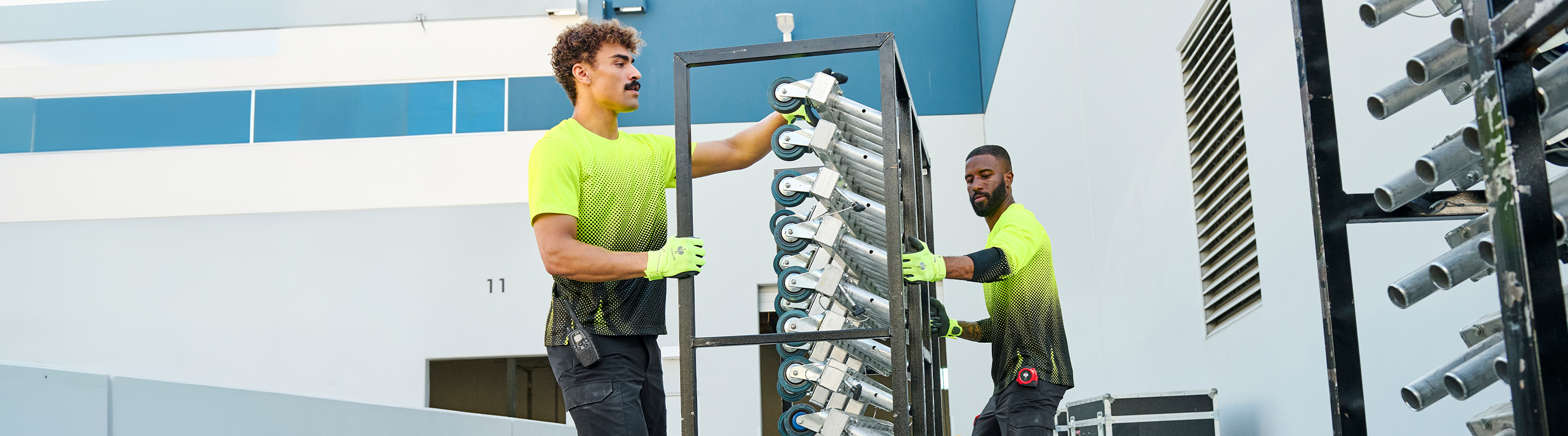 Two workers in neon yellow-green shirts and gloves install a tall rack of white cylindrical pool or spa filters against a blue and white building exterior.