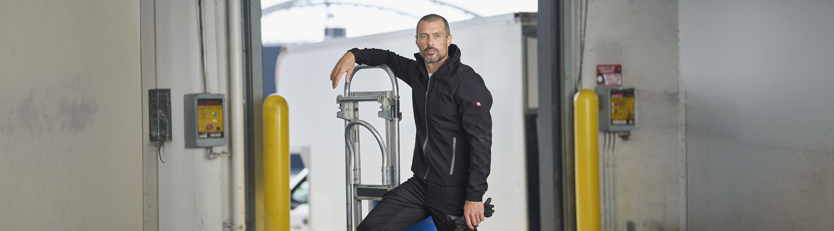 A man in a black jacket and pants leans on a cart in an industrial loading dock area, with yellow safety posts and control panels visible on either side.