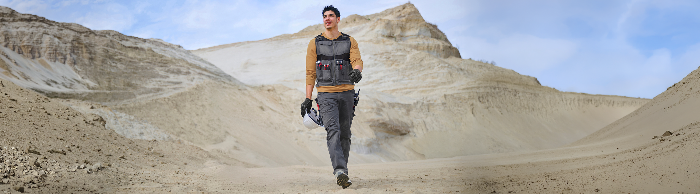 A man wearing a black and brown technical outdoor vest and dark pants walks through a sandy desert landscape with layered rock formations under a blue sky.