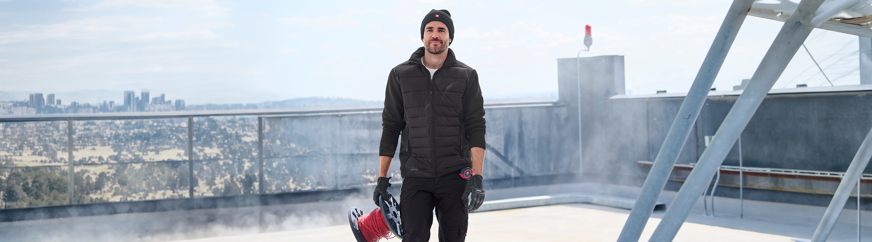 A man wearing a black puffer jacket, black beanie, and black gloves stands on a rooftop holding a red and black leaf blower, with a city skyline in the background.