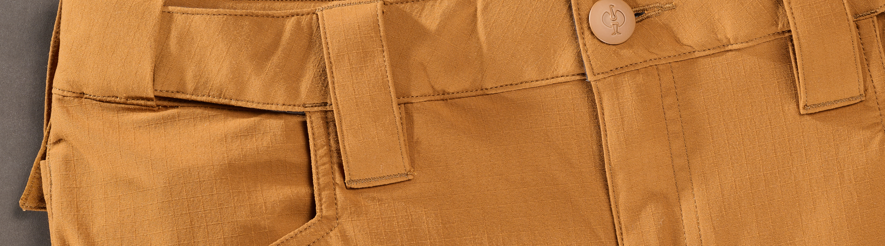 Close-up of tan or brown pants showing the waistband, belt loops, button, partial front pocket, and detailed stitching.