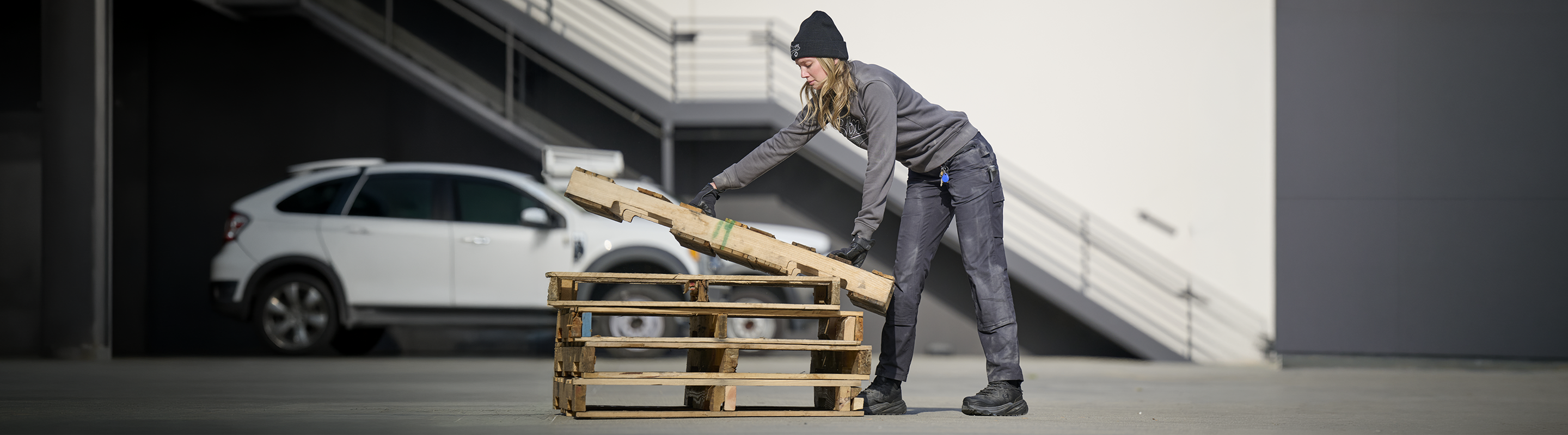 A person wearing gray work clothes and a black beanie lifts wooden boards from a stack of pallets outdoors. A white car and a staircase are visible in the background.