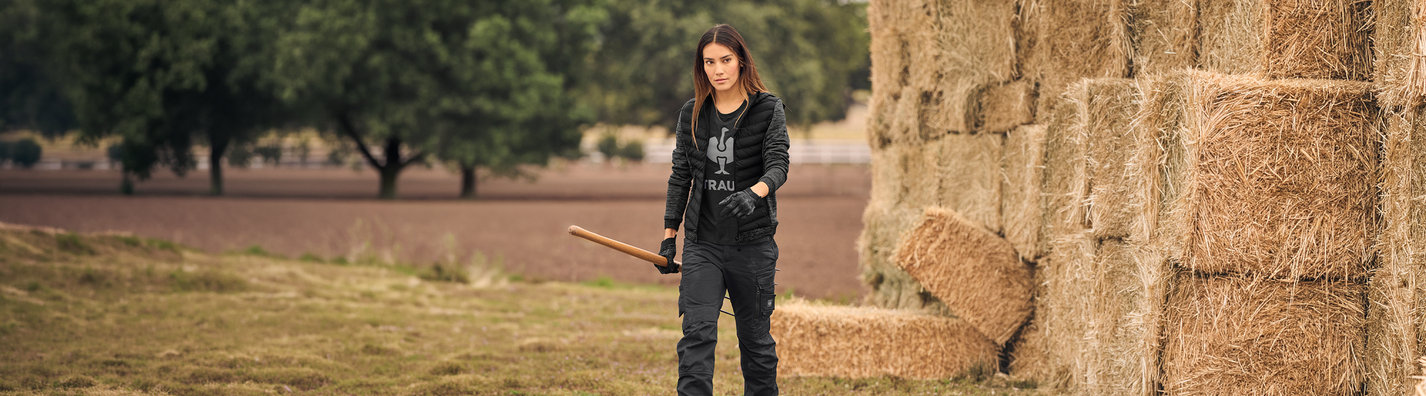A woman stands outdoors beside a large stack of hay bales, holding a wooden bat. She is wearing a black graphic T-shirt, black jacket, and black pants.