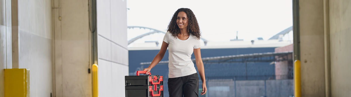 A woman with curly hair stands in a doorway holding a black suitcase with a red and black patterned handle, wearing a white t-shirt and dark pants.