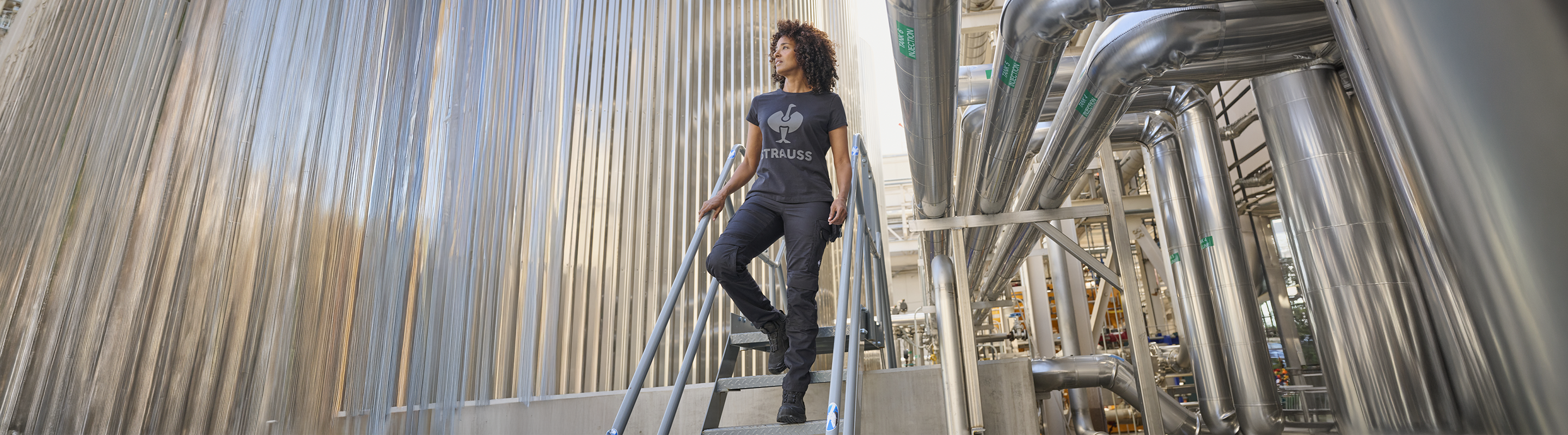 A person with curly hair wearing a TAURAS t-shirt stands on metal stairs in an industrial facility with large pipes and tanks.