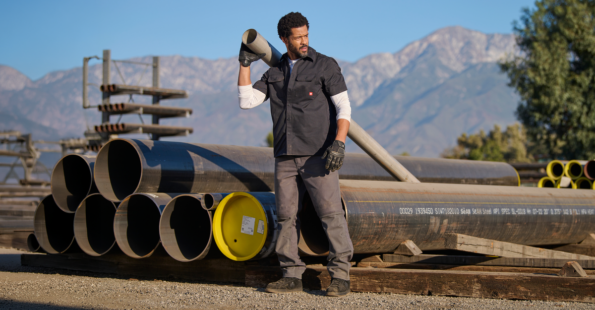 A man in a black work vest and gray pants stands outdoors in a construction yard holding a metal pipe, with large steel pipes, mountains, and trees in the background.