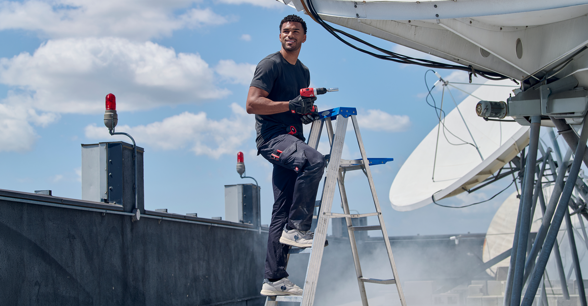 A man stands on a ladder with a power drill, working on satellite dishes on a rooftop under a blue sky with clouds.