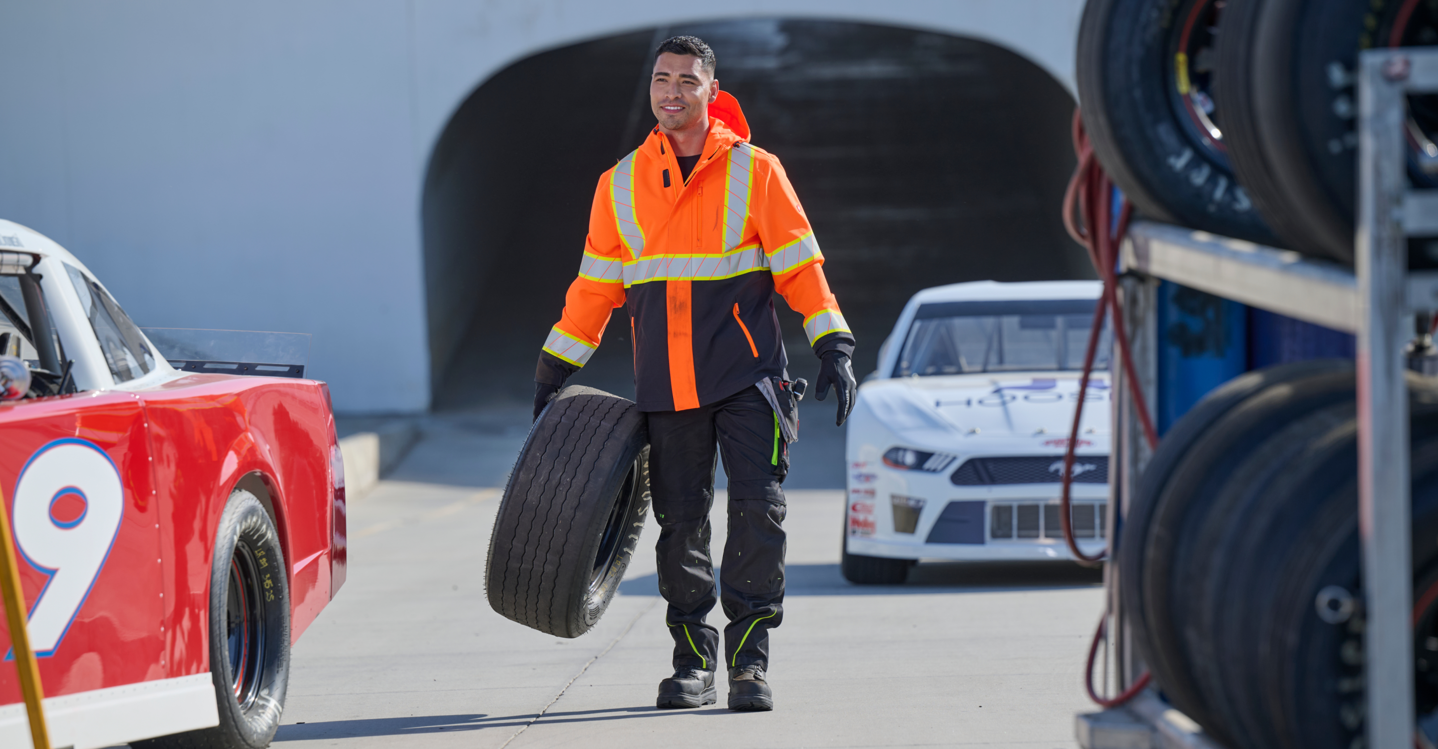 A man in a high-visibility orange and black work jacket with reflective stripes walks while carrying a large black tire on an outdoor paved area, with a white race car partially visible in the background.