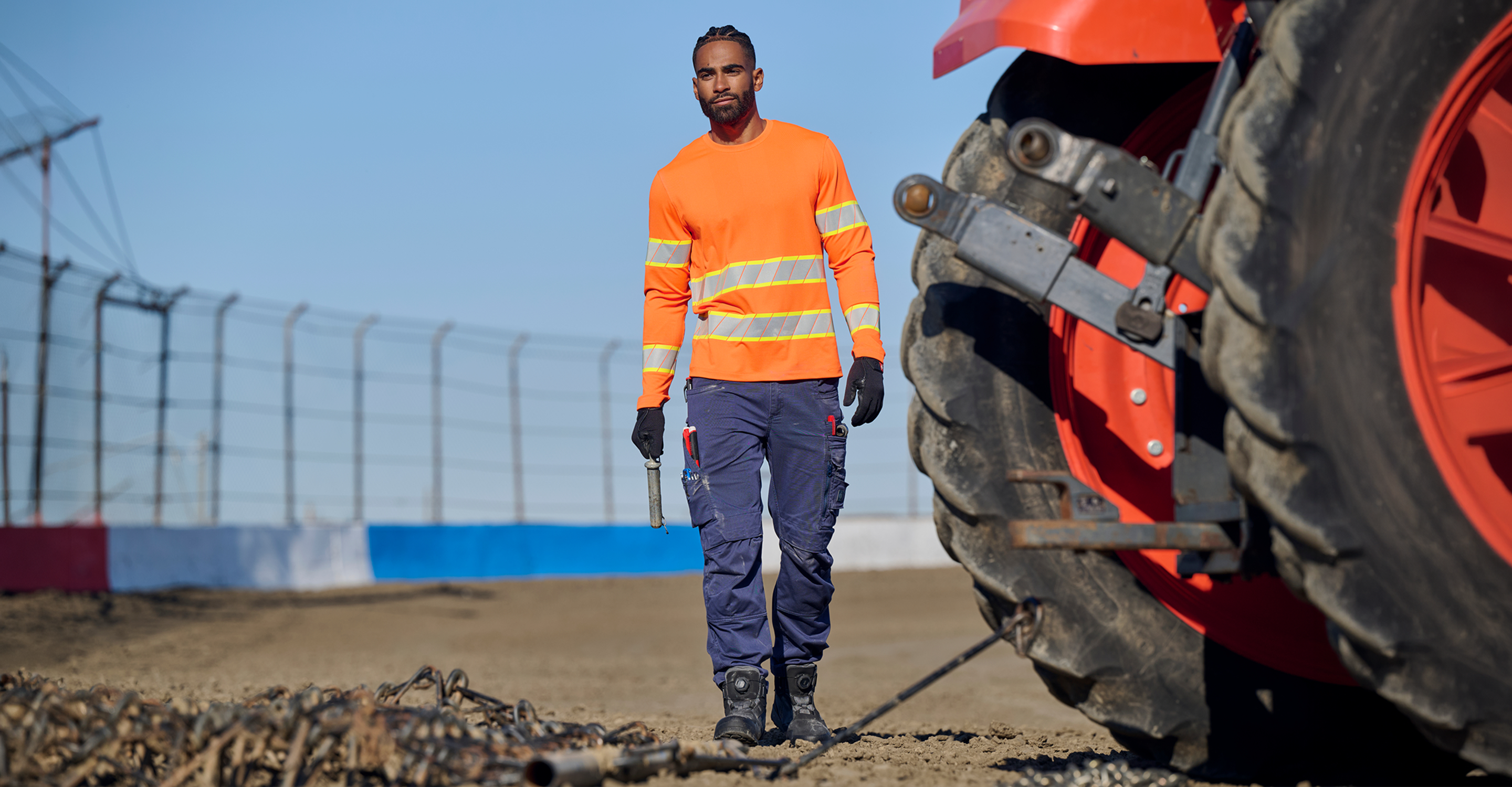 A man in a high-visibility orange and yellow safety shirt, navy work pants, and black gloves walks toward the camera in an outdoor industrial setting next to a large red tractor tire.