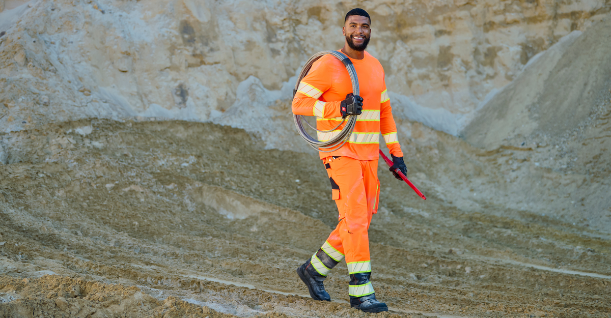 A construction worker in bright orange safety gear and reflective stripes smiles while carrying coiled cable and a red tool, walking on a dirt construction site.