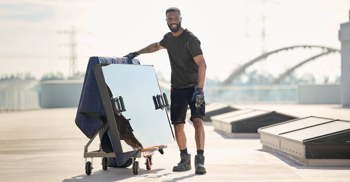 A man in work gloves moves a large, reflective glass panel on a wheeled cart outdoors. He is wearing a black shirt, shorts, and boots, with a blue padded blanket protecting the glass.