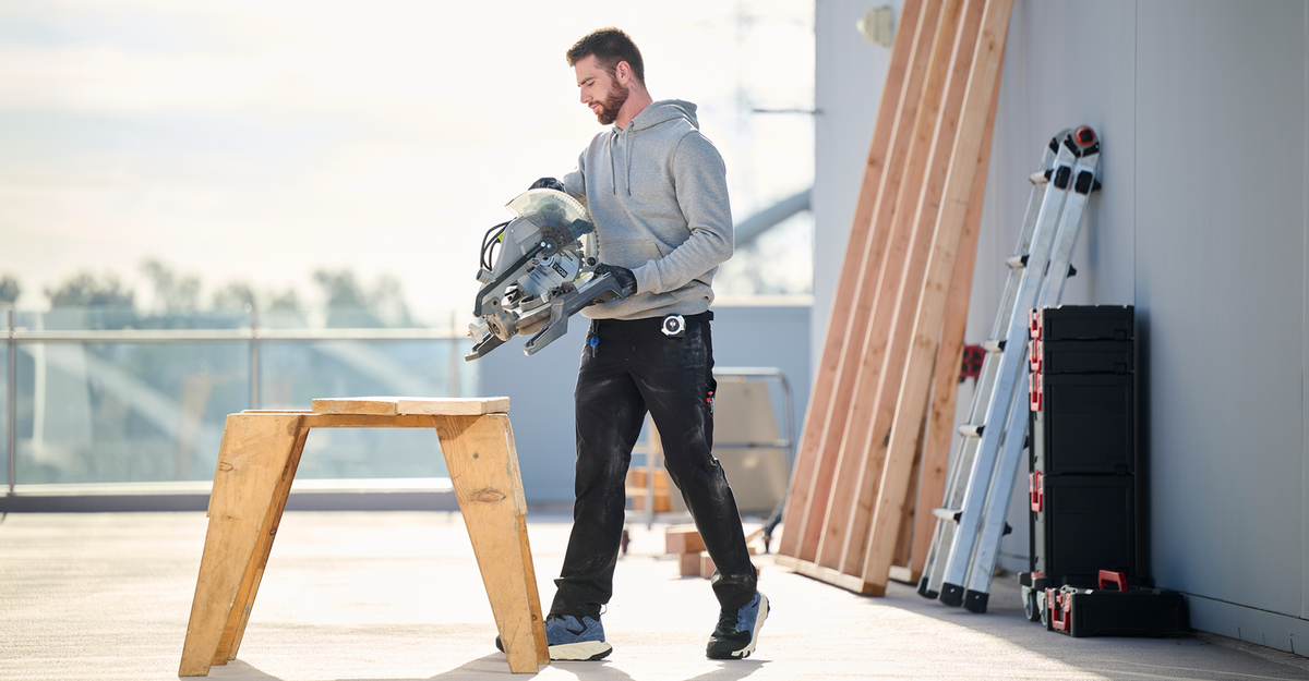 A man in a gray hoodie and black work pants carrying a circular saw stands on a concrete construction site next to a wooden saw stand, with ladders and materials in the background.