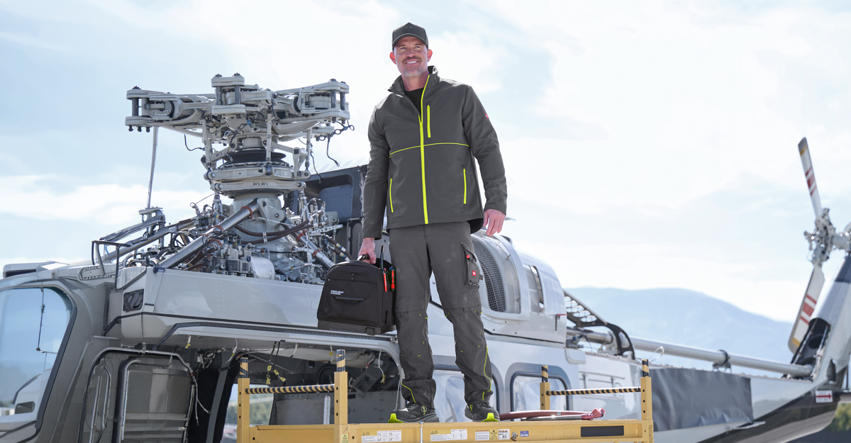 A man wearing grey workwear with yellow high-vis elements. He is standing next to a helicopter and holds a tool box in his hand.