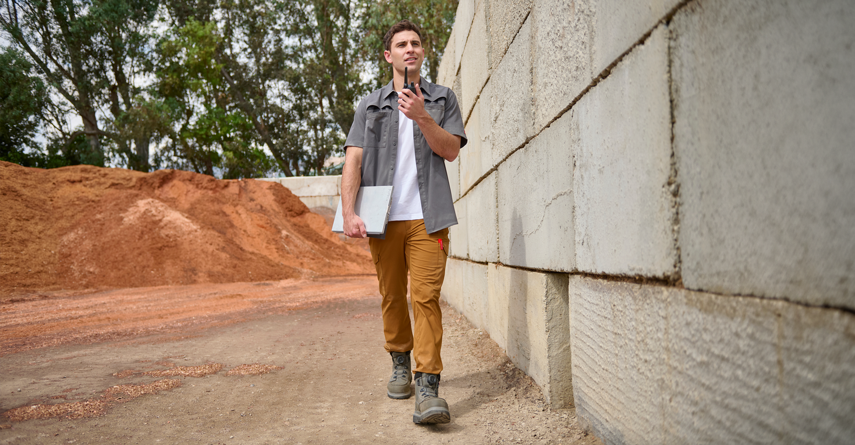 A man in work clothes walks outdoors by a concrete wall, holding a walkie-talkie and a folder. There are dirt piles and trees in the background, suggesting a construction or industrial site.
